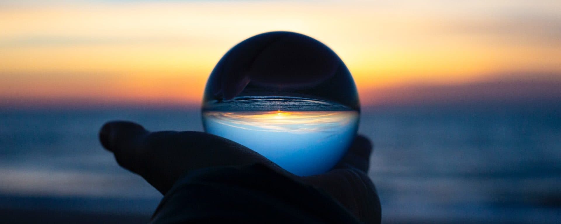 Hand holding a glass sphere that refracts an ocean sunset