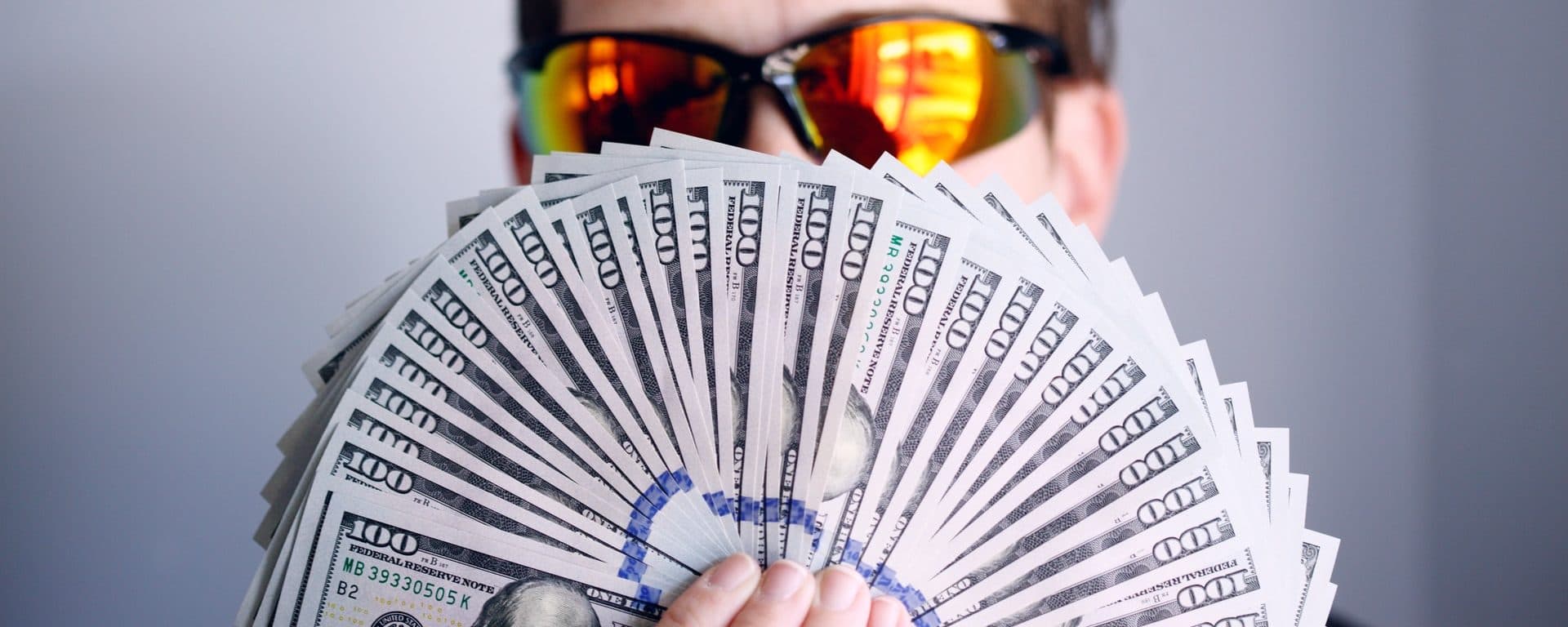 Person wearing sunglasses holding a fan of $100 bills